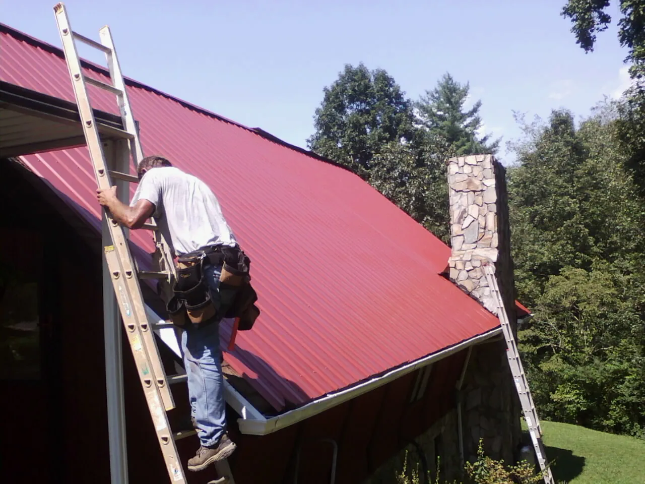 Roofer on a ladder beside a red metal roof and stone chimney — Soffit & Fascia Repair by Roofing Brothers in Astor